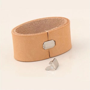 May include: A light brown leather strap with a silver metal buckle. The strap is designed to be used as a wrist strap for a camera.