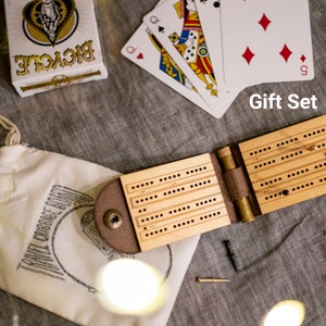 Open leather and wood travel cribbage board with cast metal person on top of a bedspread with a carrying pouch and a deck of cards, which is the "Gift Set"