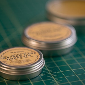 May include: Three small round metal tins with lids. The tin in the foreground is labeled "Walnut All-Natural Leather Care Dressing" and has a yellow label with black text. The other two tins are out of focus and have yellow labels.