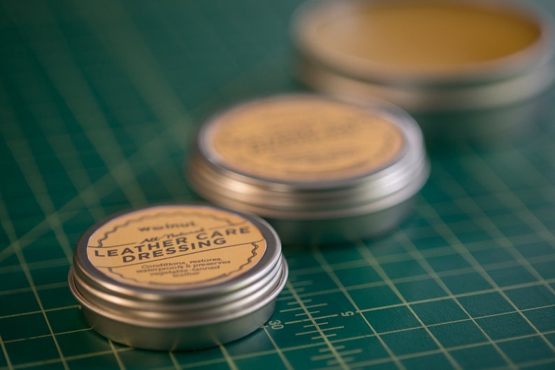 May include: Three small round metal tins with lids. The tin in the foreground is labeled "Walnut All-Natural Leather Care Dressing" and has a yellow label with black text. The other two tins are out of focus and have yellow labels.