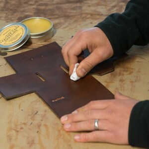 May include: A person is applying leather dressing to a brown leather piece with a white cloth. The leather piece has a cut-out design and the text "remarkable" is stamped on it. A tin of leather dressing is visible in the background.