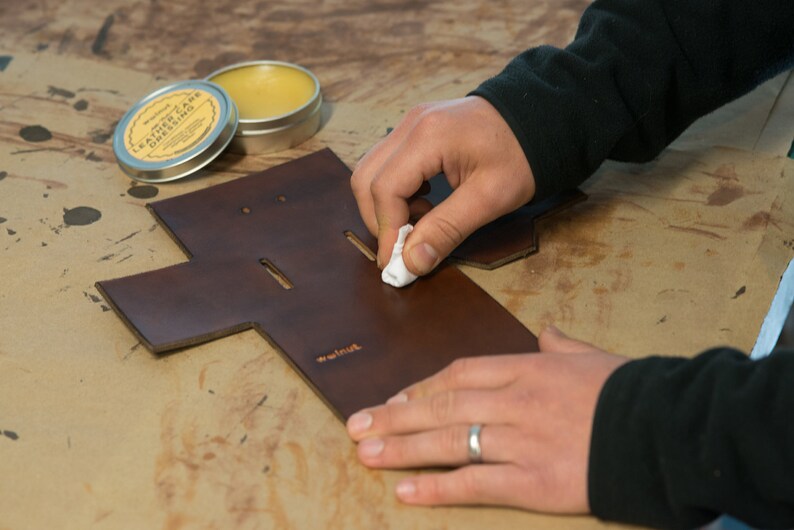 May include: A person is applying leather dressing to a brown leather piece with a white cloth. The leather piece has a cut-out design and the text "remarkable" is stamped on it. A tin of leather dressing is visible in the background.