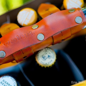 A close-up image of the honey leather variant of the 6-pack frame cinch. The beer carrier is attached to the top tube of a bike frame and is holding a yellow beer carton with six glass bottles. The cinch is embossed with the phrase walnut.