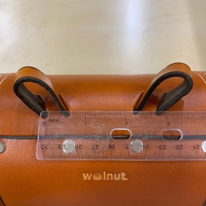 Honey leather variant of bicycle barrel bag being measured between two connecting points with clear ruler. The ruler shows that the difference between the two leather loops is three inches.