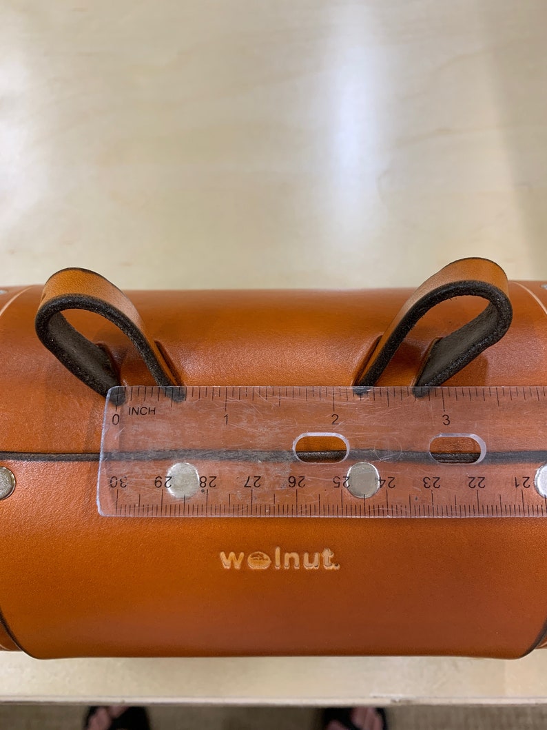 Honey leather variant of bicycle barrel bag being measured between two connecting points with clear ruler. The ruler shows that the difference between the two leather loops is three inches.