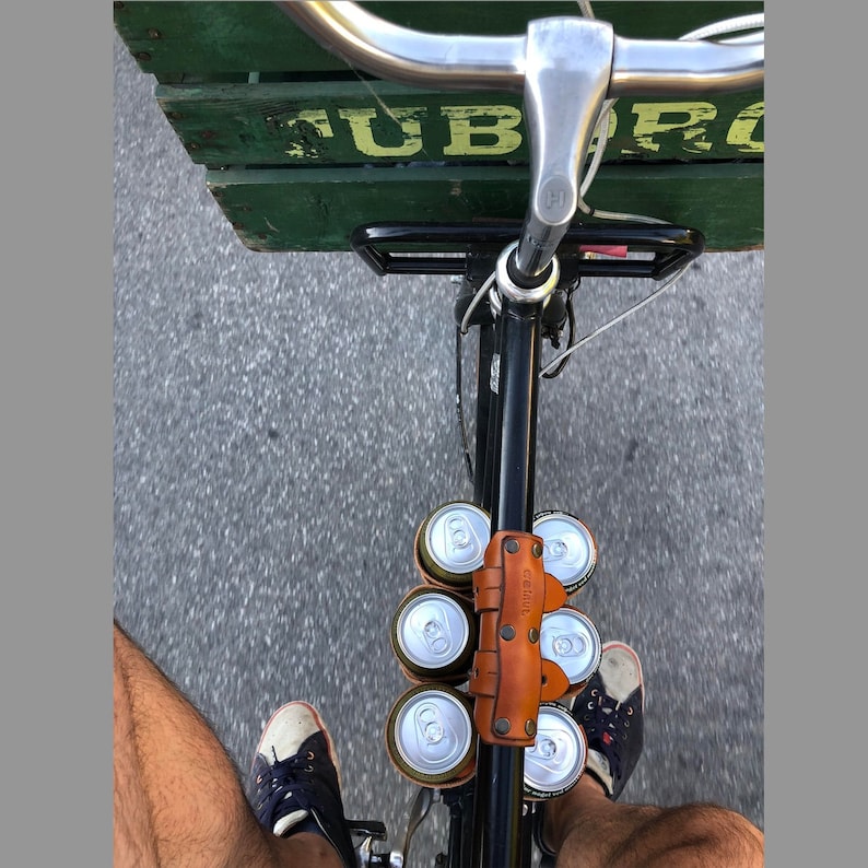 A person looking down at the top tube of a bicycle while riding it, from the saddle. Their feet are on the pedals and the ground is blurry beneath them, and there is a 6-pack of beer strapped to the top tube with a bicycle beer carrier strap