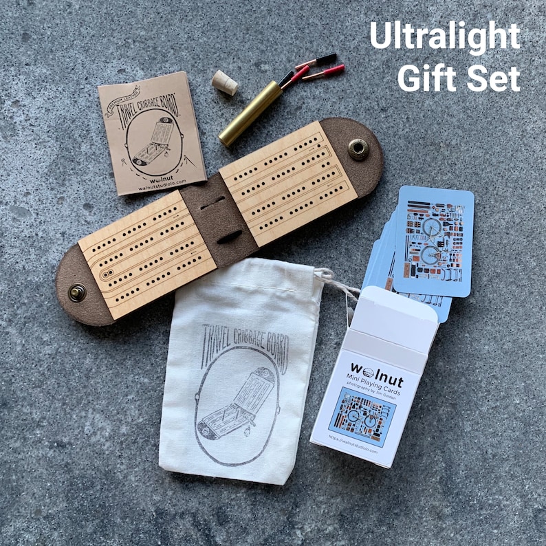 Open leather and wood travel cribbage board with wire metal pegs on a tabletop with a carrying pouch and a half-sized deck of cards, which is the "Ultralight Gift Set"