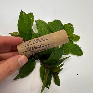 May include: A brown lip balm tube with the text "LIP BALM. Beeswax-Peppermint" is surrounded by fresh green mint leaves. The balm is held by a hand, and the background is a plain white surface. The balm is a natural skincare product.