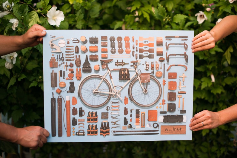 Four hands holding up an unframed bicycle poster with a variety of leather parts accessories