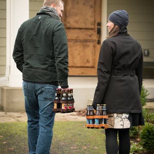 A man and a woman holding two different 6-packs of beer and chatting as they walk to the front door for a party. Each reusable leather and wood 6-pack has a different color leather, honey and dark brown