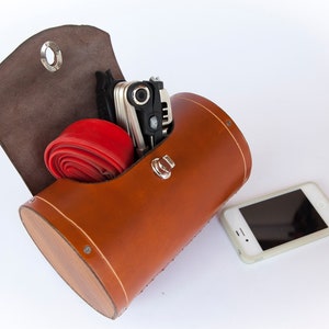 Honey leather variant of leather bicycle barrel bag on a plain white background. Inside the bag is a multi-tool and spare bike tube. An iPhone 4 is beside the bag for scale.