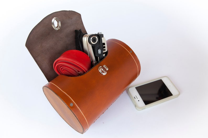 Honey leather variant of leather bicycle barrel bag on a plain white background. Inside the bag is a multi-tool and spare bike tube. An iPhone 4 is beside the bag for scale.