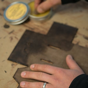 May include: A person's hand wearing a silver ring applies a yellow wax to a brown leather piece. The leather piece has a cut-out design.
