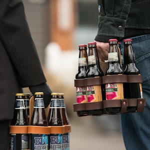 A man and a woman each holding a 6-pack of beer in a handsome leather and wood reusable 6-pack carrier, with different beers and different leather colors.