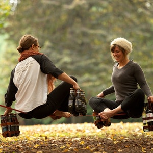 Two women balancing on a slackline in a park in Portland Oregon brewery town, holding leather 6-packs of beer as balance weights