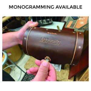 Dark brown leather bicycle handlebar bag being locked. Bag has debossing that reads "AG72217." Text above the image reads "Monogramming Available."