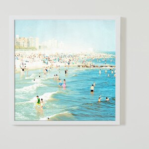 May include: A photograph of a crowded beach with people swimming and sunbathing in the ocean. The water is a light blue color and the sand is white. There are buildings in the distance.