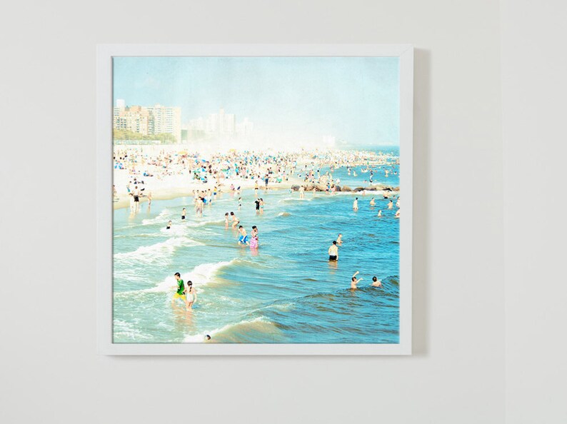 May include: A photograph of a crowded beach with people swimming and sunbathing in the ocean. The water is a light blue color and the sand is white. There are buildings in the distance.