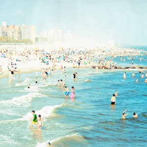 May include: A crowded beach scene with people swimming and sunbathing in the water. The beach is sandy and there are buildings in the background.