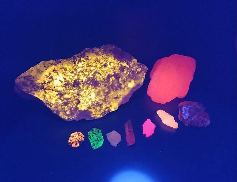 May include: A collection of fluorescent minerals under UV light. The largest piece is a yellow-green rock with a rough texture. Other pieces include a red, a green, a pink, a white, and a purple mineral.