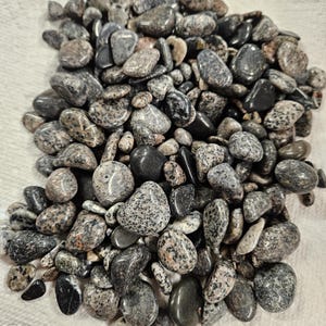 May include: A collection of smooth, polished stones in various shades of gray, black, and speckled patterns. The stones vary in size and shape, with some appearing glossy and others matte. The natural stones are arranged on a white surface.