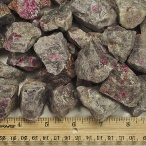 May include: A collection of rough, uncut ruby stones. The stones are a mix of gray and tan, with vibrant red inclusions. The rocks vary in size and shape, creating a natural, raw aesthetic. A ruler is visible in the image.