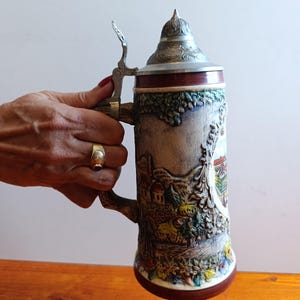 Op de afbeelding: Een hoge keramische bierpul met een gedetailleerd reliëflandschap met huizen en bomen, bekroond met een puntig metalen deksel. De pul heeft een stevig handvat en een donkerbruine basis met ingewikkelde gegoten texturen en accenten.