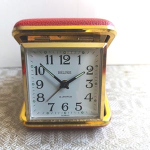 Könnte beinhalten: Eine Vintage-Reiseuhr mit goldfarbenem Rahmen und rotem Kunstlederbezug. Das Zifferblatt ist weiß mit schwarzen Ziffern und Zeigern, und die Wörter "DELUXE" und "2 JEWELS" sind auf dem Zifferblatt aufgedruckt. Die Uhr ist unten mit "GERMANY" gekennzeichnet.