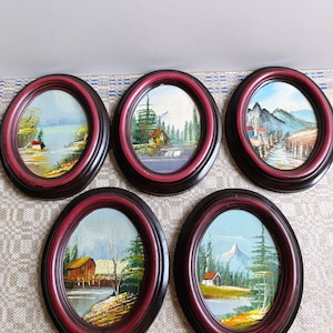 May include: Five small oval framed landscape paintings. Each painting depicts a different scene with a house, trees, and water. The frames are dark brown with a red inner edge. The paintings are in a variety of colors.