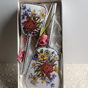Könnte beinhalten: Ein Vintage-Kosmetikset mit Handspiegel, Bürste und Kamm, präsentiert in einer Box. Das Set hat ein Blumendesign mit roten, lila, gelben und weißen Blumen. Die Griffe sind silberfarben.