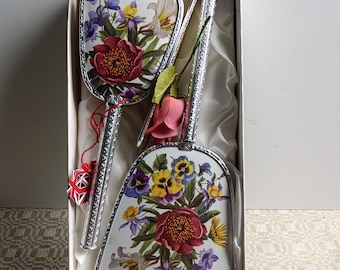 Puede incluir: Un juego de tocador vintage con espejo de mano, cepillo y peine, presentado en una caja. El juego tiene un diseño floral con flores rojas, moradas, amarillas y blancas. Los mangos son plateados.