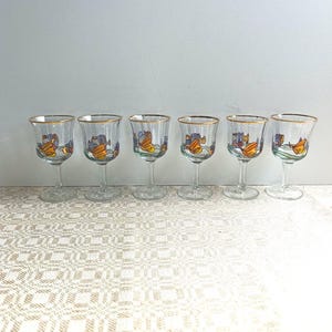 Könnte beinhalten: Sechs klare Weingläser mit goldenen Rändern. Jedes Glas hat ein farbenfrohes Design eines Bootes mit orangefarbenen Segeln und blauen und grünen Akzenten. Die Gläser sind in einer Reihe auf einer gemusterten Oberfläche angeordnet.