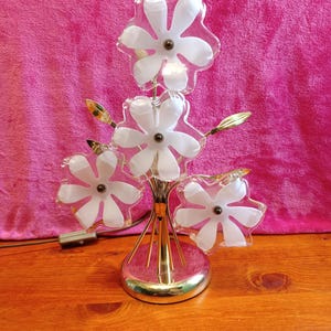 Può includere: Una lampada da tavolo floreale vintage con base e stelo color oro. La lampada presenta cinque paralumi a forma di fiore bianco e rosa. La lampada è su una superficie di legno con sfondo rosa.