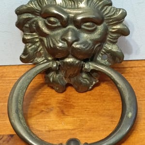 Könnte beinhalten: Antiker Türklopfer aus Messing in Form eines Löwenkopfes mit rundem Griff. Das Gesicht des Löwen ist detailliert mit Mähne, Augen, Nase und Mund. Der Griff hat einen Durchmesser von etwa 10 cm.