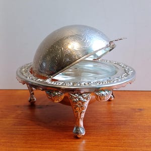 Pode incluir: Manteigueira prateada com tampa abobadada, aberta para revelar um prato de vidro. O prato assenta em pernas ornamentadas com detalhes florais. A tampa e a base apresentam padrões gravados intrincados, adicionando um toque de elegância.