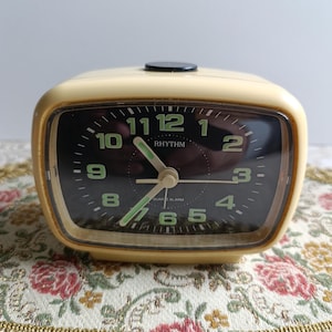 May include: A cream-coloured square alarm clock with a black face and green numbers. The clock says "RHYTHM" and "QUARTZ ALARM".