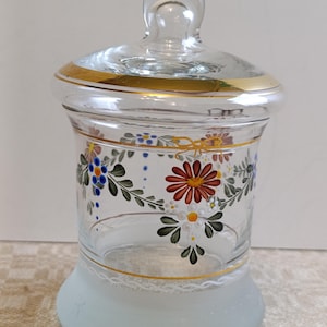 May include: A clear glass apothecary jar with a lid, featuring hand-painted floral designs in red, blue, and yellow. The jar has a gold rim and a frosted base, creating an elegant vintage aesthetic.