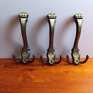 Puede incluir: Tres percheros de estilo antiguo con acabado bronce y dorado. Cada gancho tiene una parte superior decorativa, un cuerpo largo y curvado y dos ganchos curvos en la parte inferior. Los ganchos están montados sobre una superficie de madera.