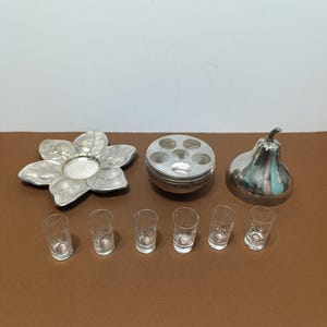 以下が含まれることがあります： 銀色のバー用品のコレクション。花型の皿、複数の開口部がある丸い容器、梨型のアイテム、6つの小さなショットグラスが含まれています。アイテムは茶色の表面に配置されています。