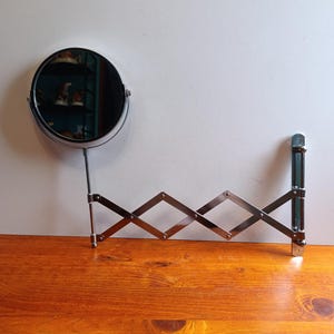 May include: A circular wall-mounted mirror with a polished chrome finish, featuring an adjustable accordion-style extending arm. The mirror is attached to a vertical mounting bracket, allowing for horizontal extension and positioning as needed.