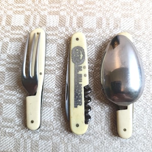 Può includere: Un set di tre utensili da viaggio vintage: una forchetta, un coltello multiuso con cavatappi e un cucchiaio. Ogni utensile ha un manico color crema e un'estremità di lavoro in acciaio inossidabile. Il coltello è contrassegnato con la scritta "M.H. BILMOSER".