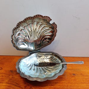 Può includere: Un piatto in metallo argentato a forma di conchiglia con coperchio incernierato che si apre verso l'alto. L'interno presenta un design a coste smerlate con una superficie riflettente. Un piccolo coltello spalmabile abbinato riposa all'interno.