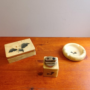 Pode incluir: Um conjunto vintage de três itens: uma caixa retangular, um isqueiro e um cinzeiro redondo. Cada peça é de cor creme com um design floral. A caixa tem uma tampa articulada. O isqueiro tem um topo de metal dourado. O cinzeiro é raso.