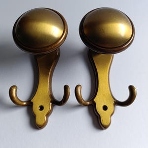 May include: Two gold-toned metal hooks with a rounded top and three hooks on each. The hooks are mounted on a white background.