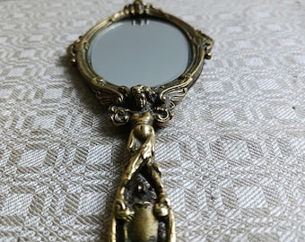May include: A vintage brass hand mirror with an oval mirror and elaborate detailing. The handle features a sculpted figure and decorative elements. The mirror is a vintage item, likely from the early 20th century.