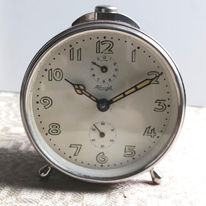 Può includere: Una sveglia vintage color argento con quadrante color crema. L'orologio presenta numeri neri, due quadranti più piccoli e il marchio "Kiengle". L'orologio ha un quadrante rotondo, una cornice in metallo e due piedini.