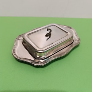 May include: A silver butter dish with a rectangular lid and a decorative handle. The dish sits on a matching scalloped tray. The handle is a small, stylized figure. The set is displayed against a green background.