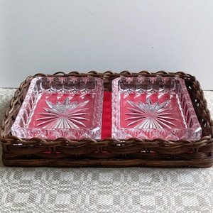 Puede incluir: Una bandeja de mimbre trenzado marrón contiene dos platos de vidrio rectangulares transparentes con diseños de estrella. Los platos descansan sobre un forro de tela roja. La bandeja está sobre una superficie estampada.