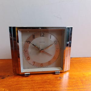 May include: A vintage silver-toned Montana clock with a square face. The clock has a light-colored face with black numerals and hands. The clock is sitting on a wooden surface.