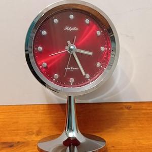 Puede incluir: Un reloj despertador Rhythm cromado con una llamativa esfera roja. El reloj tiene una esfera redonda con manecillas e indicadores de hora plateados, y el texto "Rhythm" se muestra encima del centro. Se apoya en una base cromada.